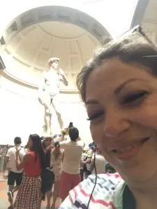 Elizabeth Macias, M4 Travel Architect, of San Antonio, Texas inside the Uffizi Gallery with a copy of Michelangelo's David statue visible, representing the artistic depth of Italian culture.